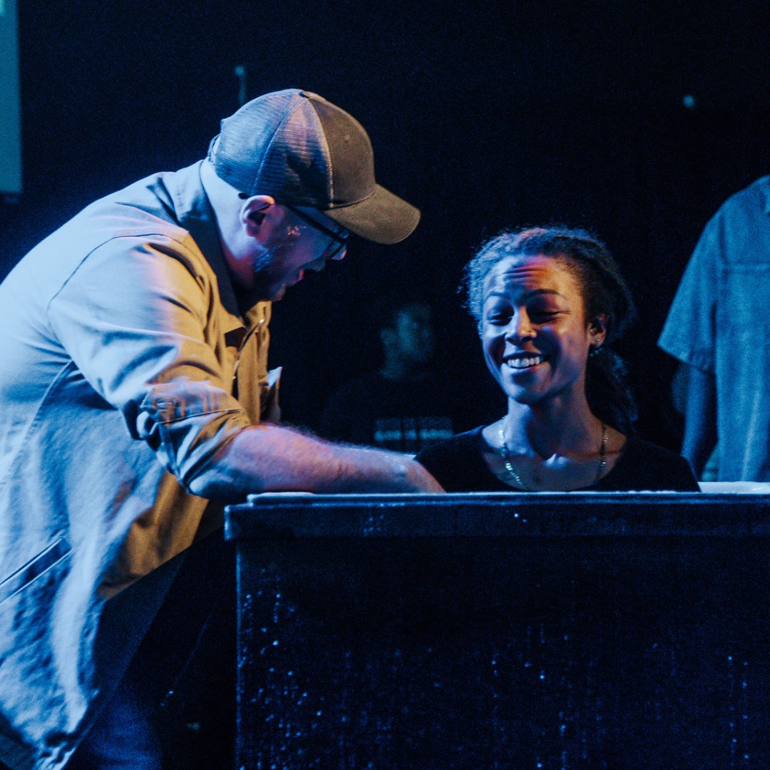 A man wearing a cap and glasses smiling and leaning over a baptism pool while a woman beside him smiles with her eyes closed.