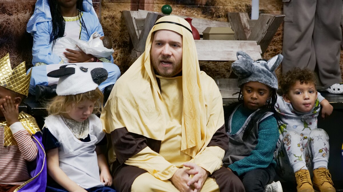 An adult dressed in a yellow and brown biblical costume sits among children in various costumes including a sheep, donkey, and princess.