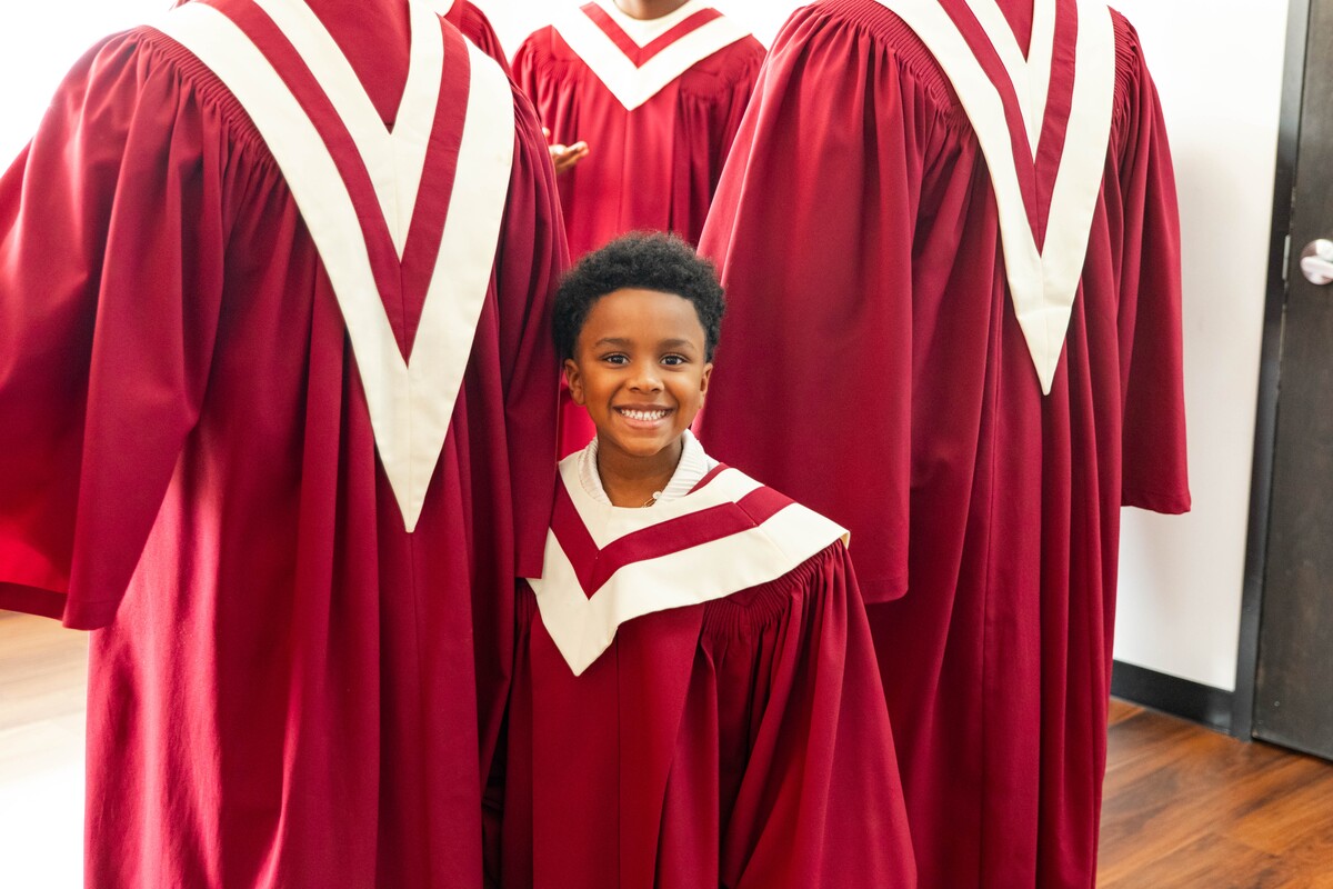Smiling young boy in red choir robe with white collar standing among adults in similar robes.