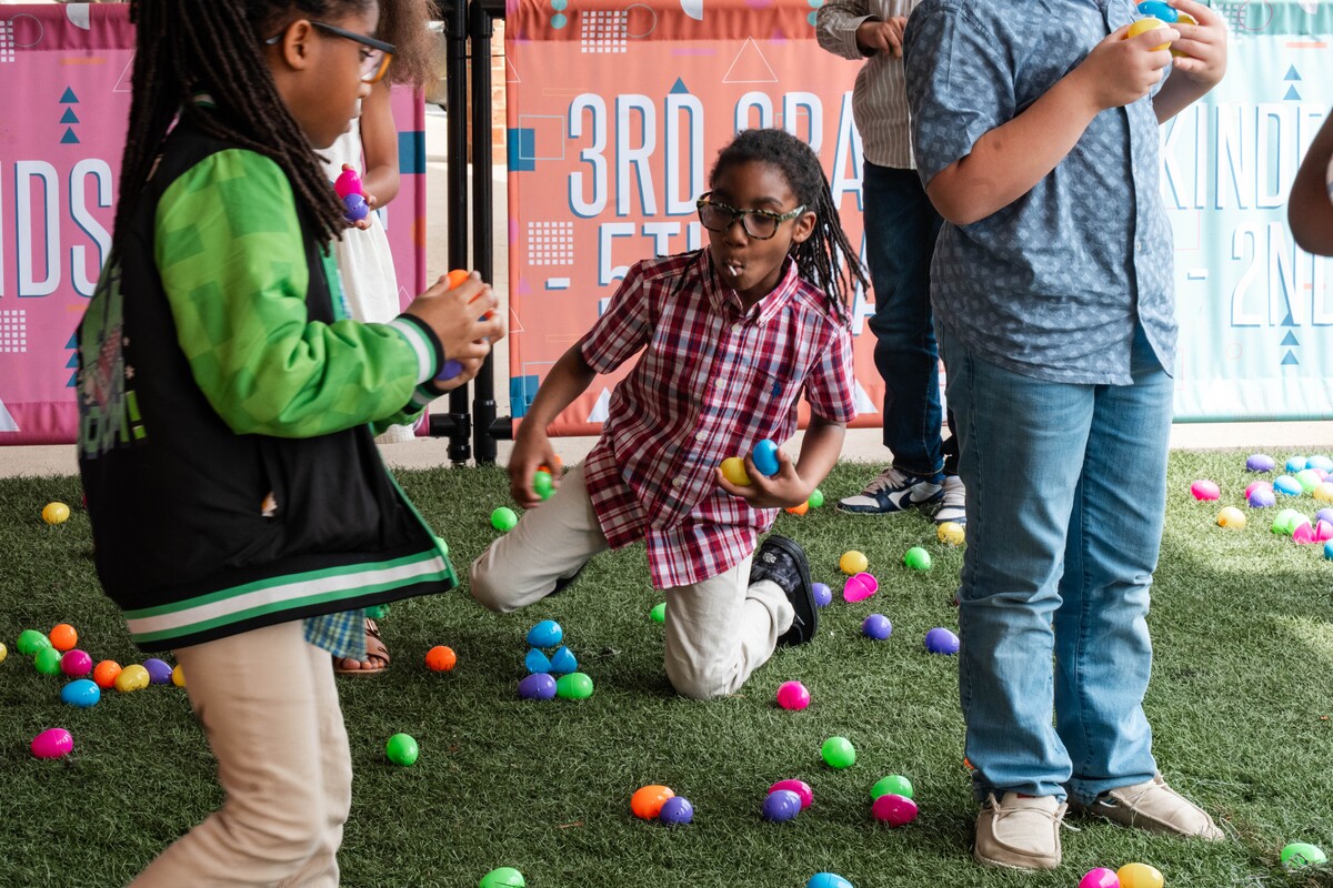 Children participating in an Easter egg hunt on green artificial grass with colorful plastic eggs scattered around.