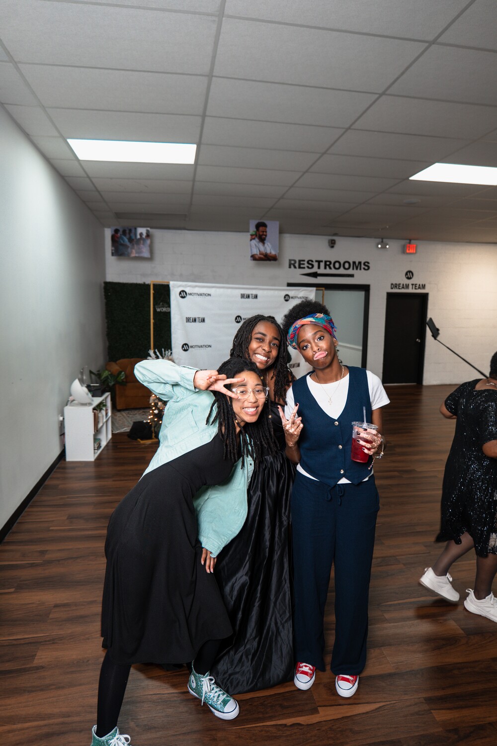 Three young women smiling and posing together indoors, one holding a drink and making a peace sign, with a wooden floor and restroom signs in the background.