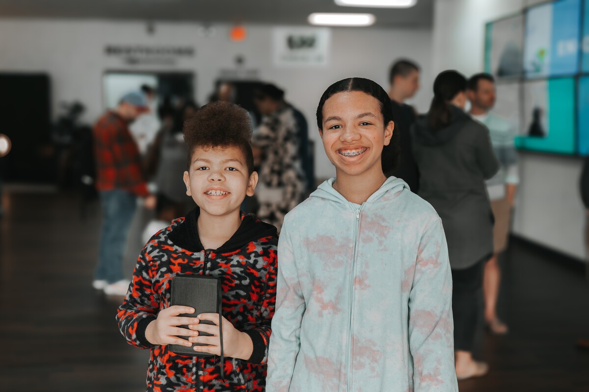 Two smiling children wearing hoodies, one holding a black book, standing in a room with several people in the background.