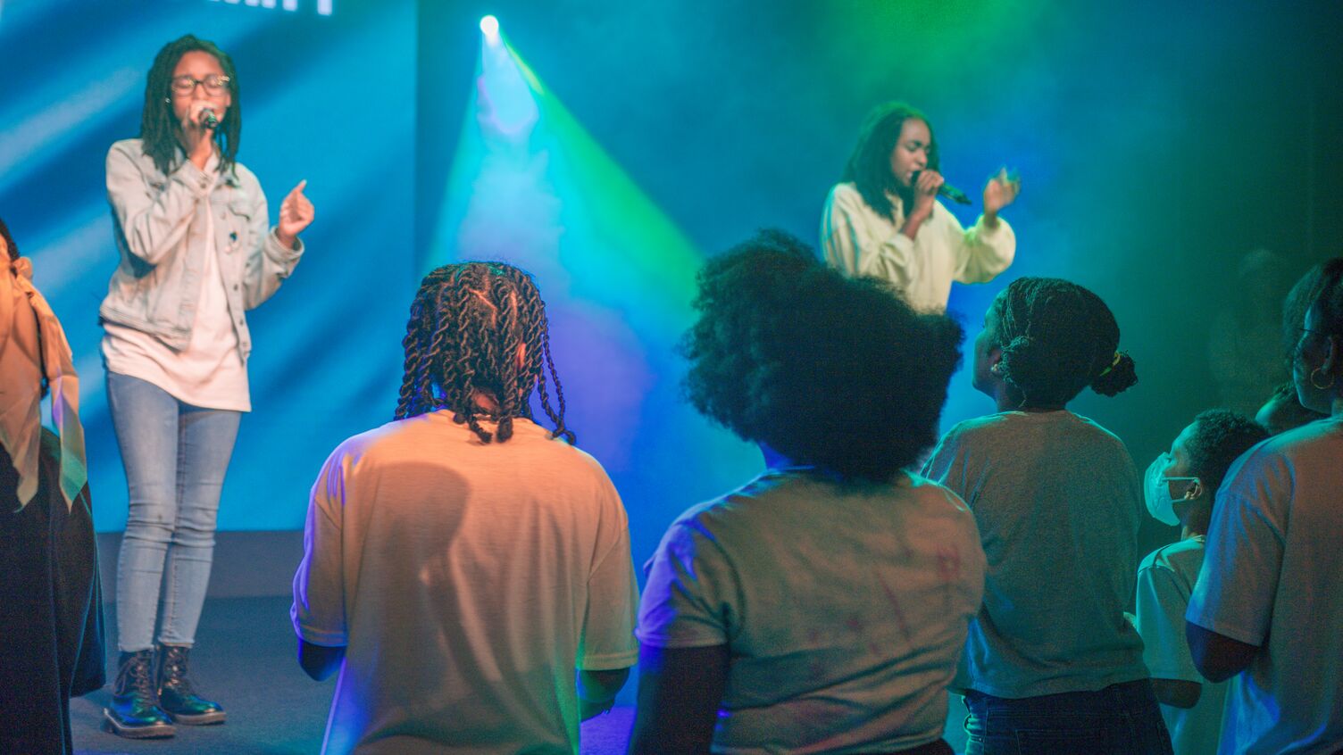 Young people standing and singing on a stage with colorful blue and green lights in the background.