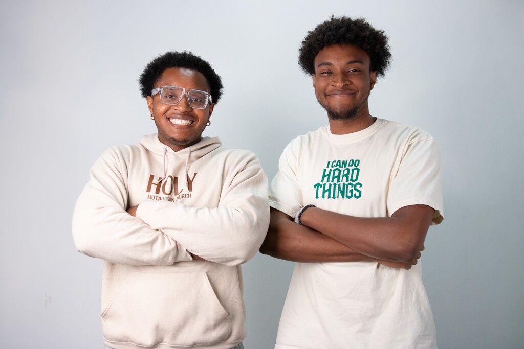 Two young men with crossed arms smiling; one wears glasses and a hoodie reading 'HOLY MOTIVATION CHURCH', the other a t-shirt saying 'I CAN DO HARD THINGS'.