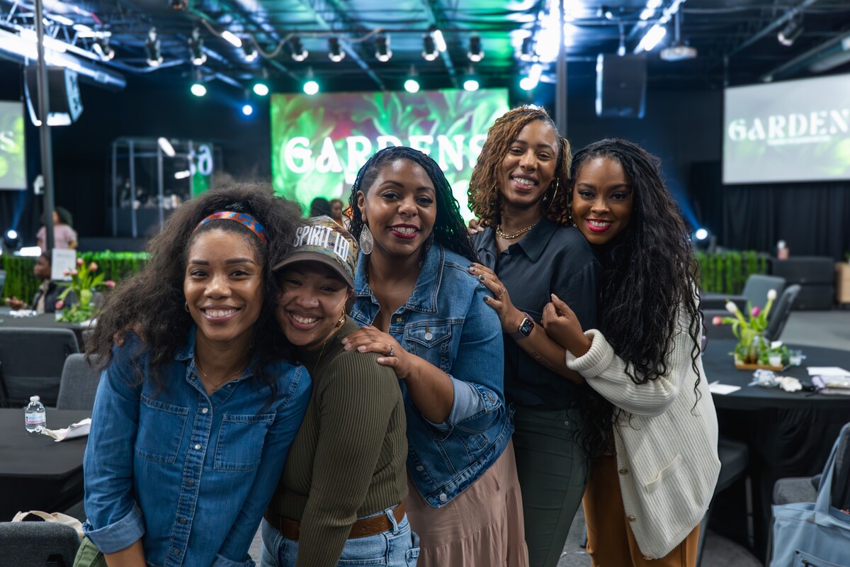 Five smiling women posing closely together in a warmly lit event space with a stage in the background.