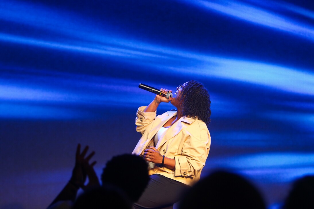 Woman with curly hair singing energetically into a microphone on stage with blue light beams in the background.
