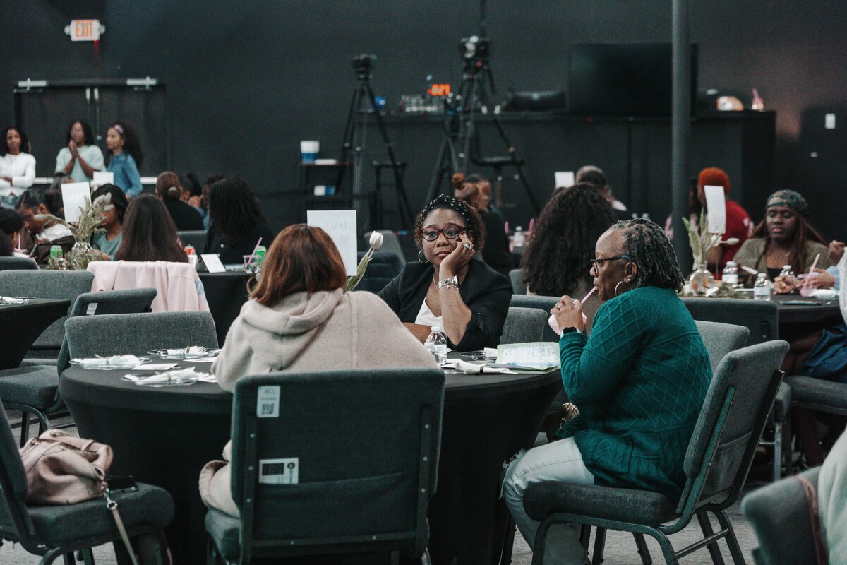 Three women sitting at a round table in a conference or event setting, one drinking from a straw and another resting her face on her hand.