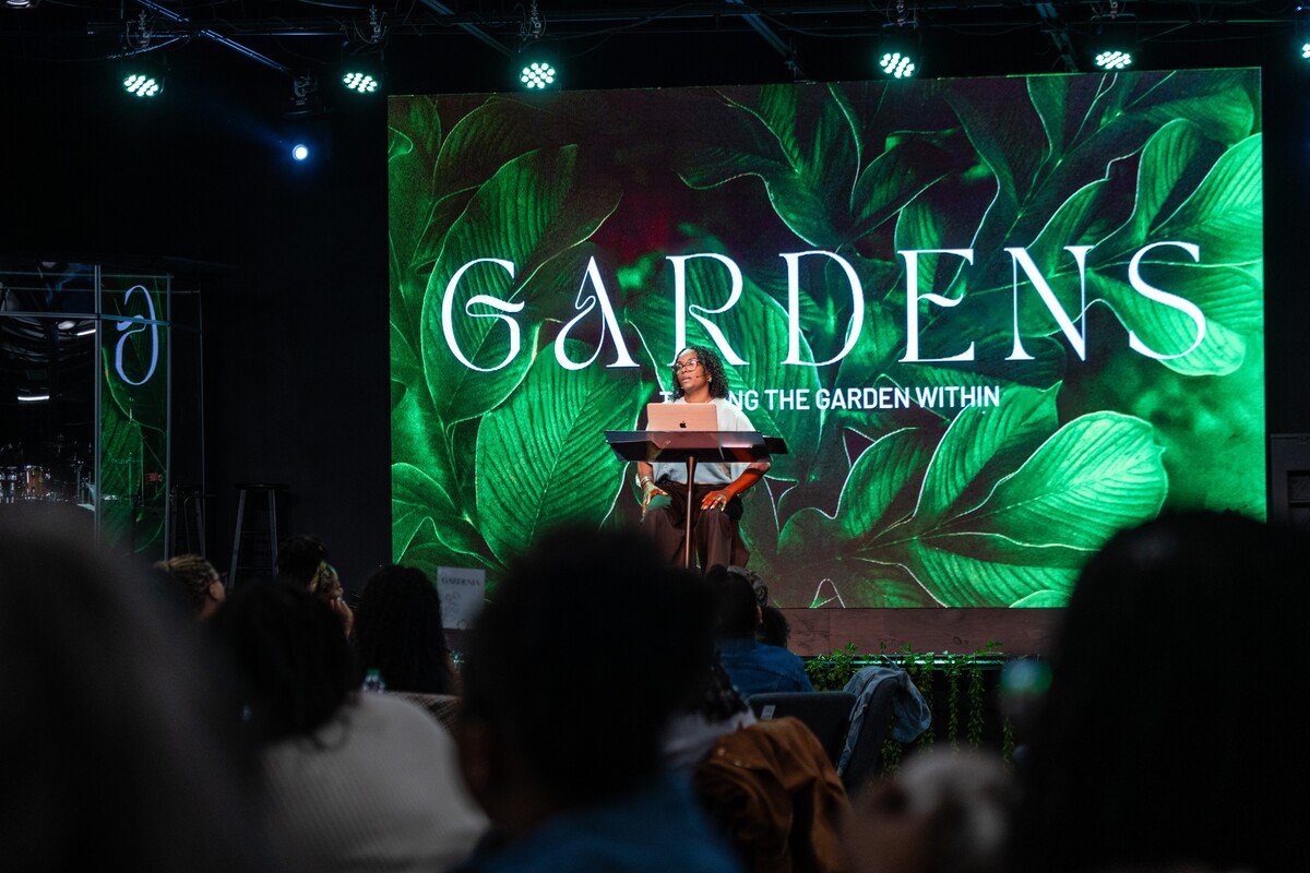 Speaker sitting at a podium with a laptop in front of a large screen displaying the word 'GARDENS' and green leafy background during an indoor event.