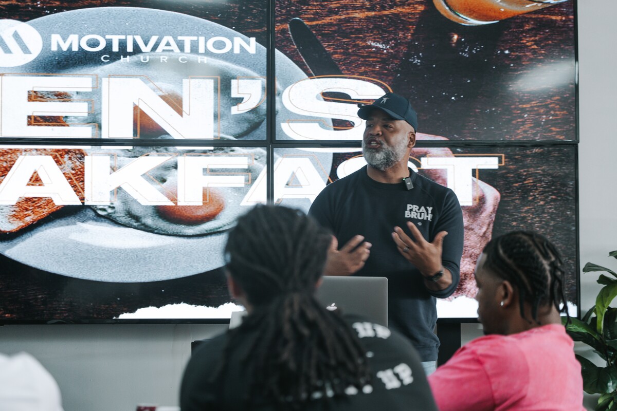 Man in a black 'PRAY BRUH' shirt speaking to a small audience with a screen behind showing 'Motivation Church Men's Breakfast'.