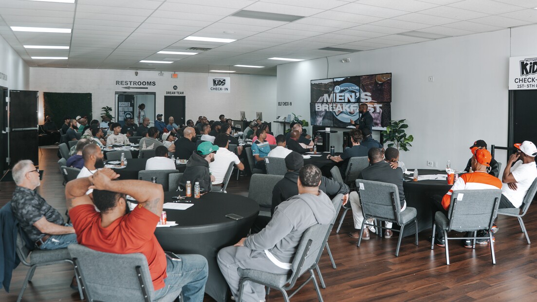 Group of men seated at round tables listening to a speaker during a men's breakfast event in a large room.