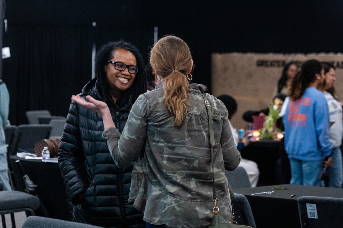 Two women engaged in conversation at an indoor event, one smiling and wearing glasses and a black jacket, the other with a ponytail and wearing a camouflage jacket.