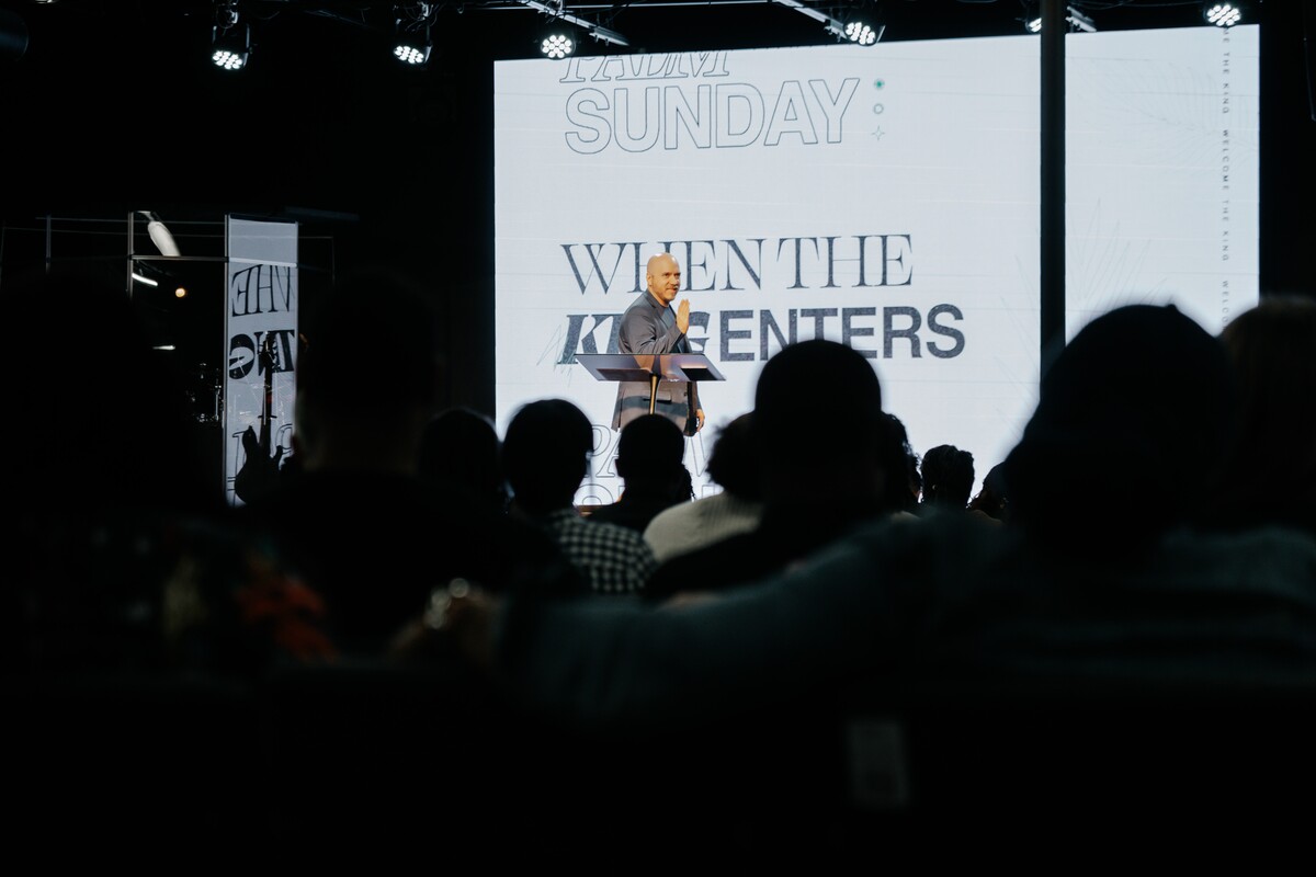 A man speaking on stage in front of an audience with a large screen behind him displaying the text 'WHEN THE KING ENTERS'.