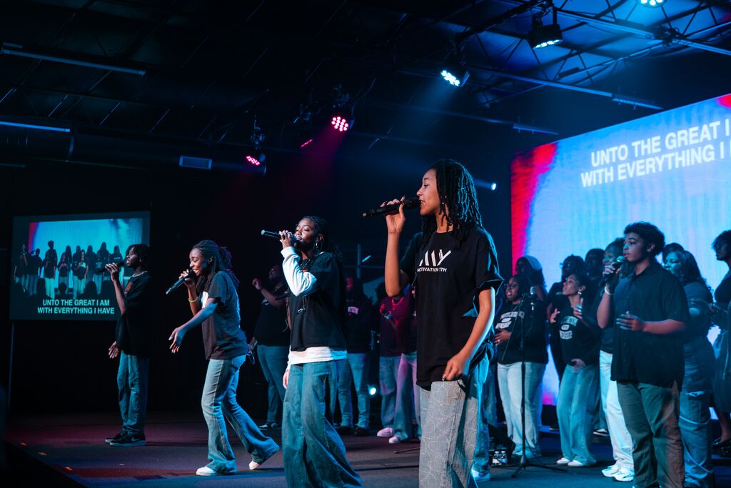 Group of young singers performing on stage with microphones and colored lights, lyrics displayed on screens behind them.