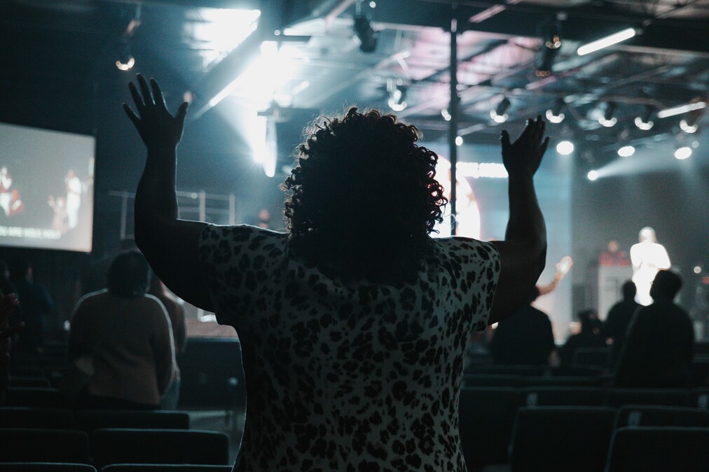 Person with raised hands in a dimly lit auditorium with stage lights and a speaker in the background.