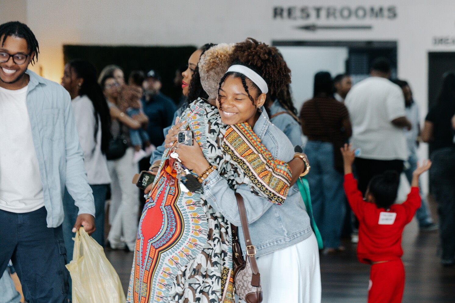 Two women warmly hugging in a busy indoor gathering with diverse people in the background.