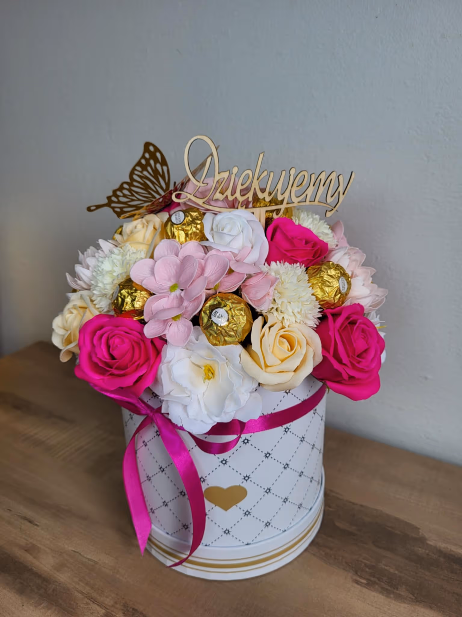 Flowerbox z różowymi, kremowymi i białymi kwiatami oraz pralinami w złotych owijkach, ozdobiony drewnianym napisem „Dziękujemy” i złotym motylem