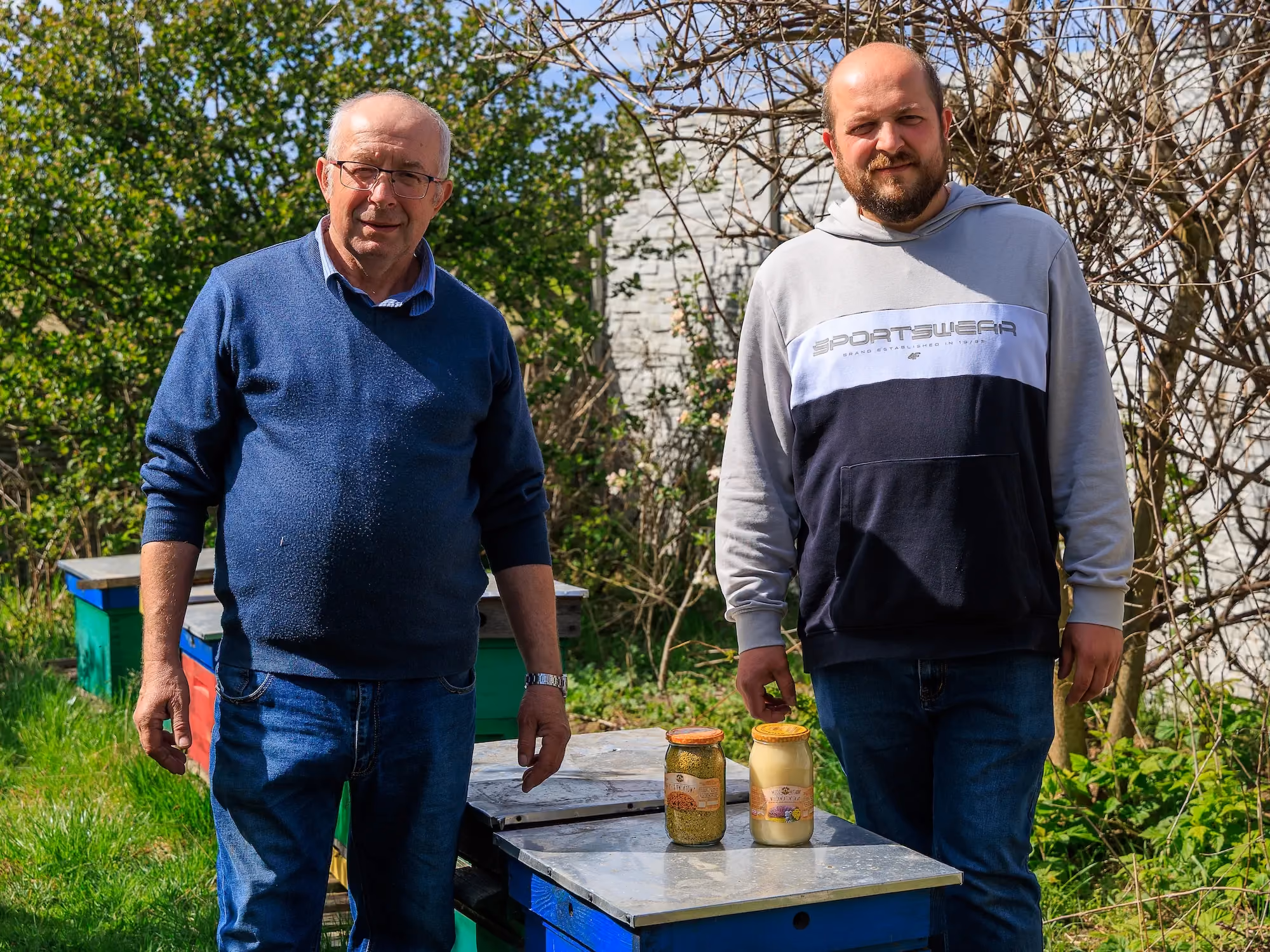 Dwaj mężczyźni stoją w pasiece przy ulach, na jednym z uli ustawione są dwa słoiki z miodem i pierzgą.