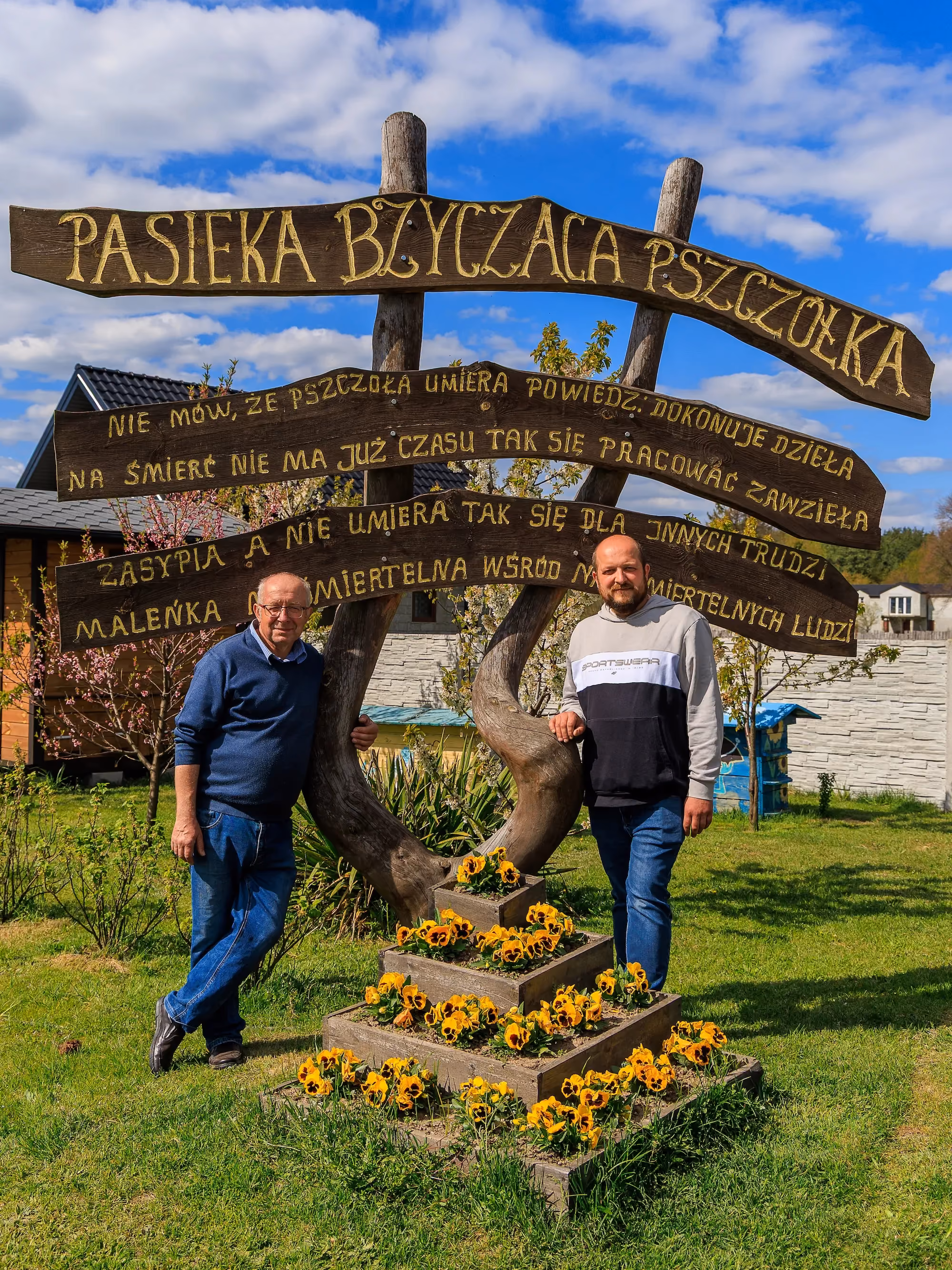 Dwaj mężczyźni przy drewnianej tablicy z nazwą „Pasieka Bzycząca Pszczółka” i wierszem, ustawionej za rabatą z żółtymi kwiatami w ogrodzie.
