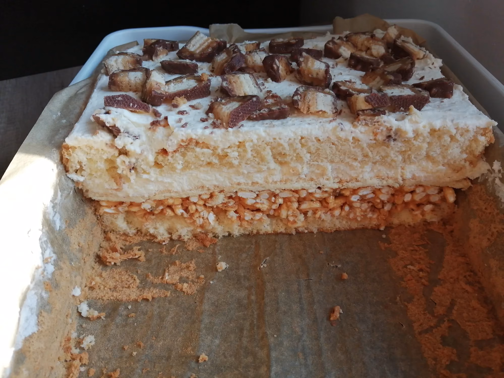 Ciasto warstwowe w formie prostokątnej, z wyraźną warstwą jasnego biszkoptu, kremu oraz chrupiącego spodu, dekorowane na wierzchu kawałkami czekoladowych batonów