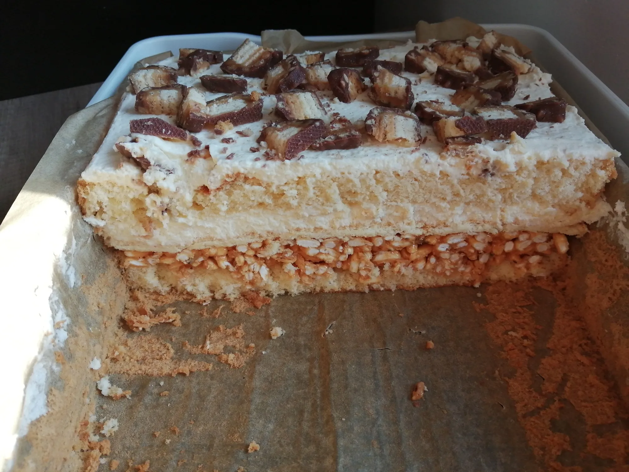 Ciasto warstwowe w formie prostokątnej, z wyraźną warstwą jasnego biszkoptu, kremu oraz chrupiącego spodu, dekorowane na wierzchu kawałkami czekoladowych batonów