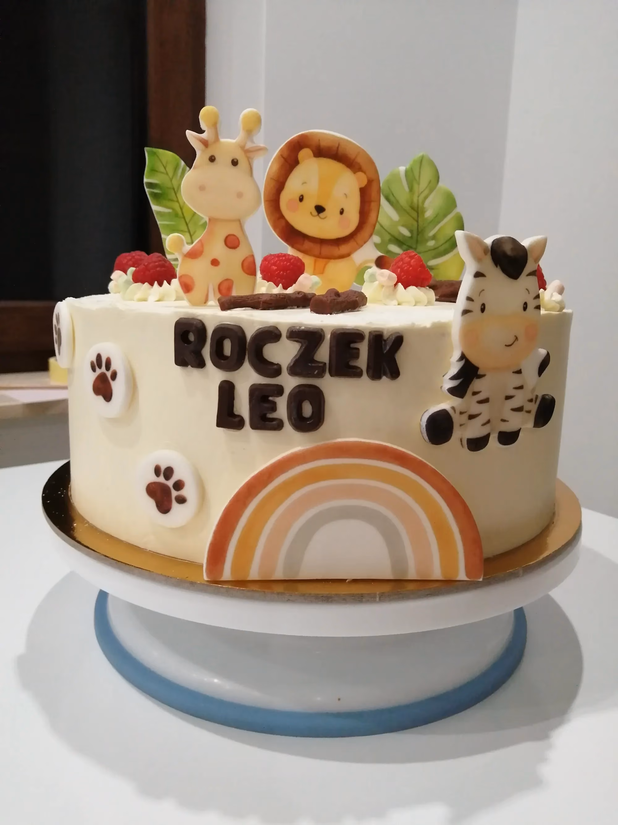 Tort z jasnym kremem, dekorowany cukrowymi zwierzętami, tęczą i napisem „ROCZEK LEO”
