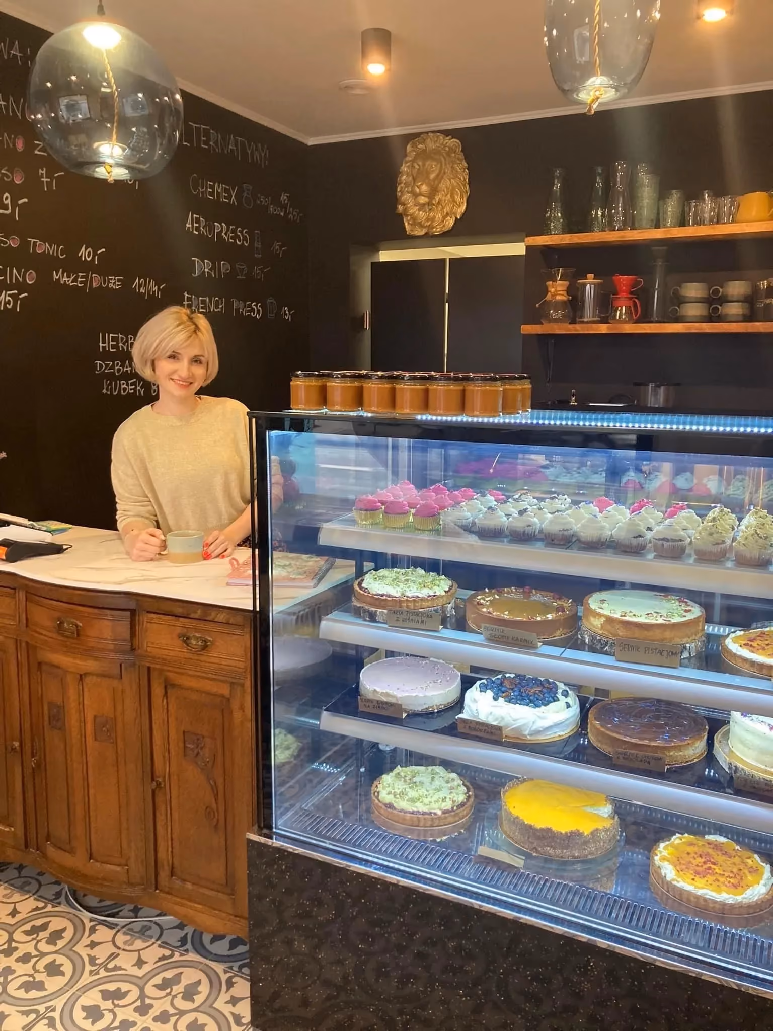 Uśmiechnięta kobieta za ladą w cukierni; obok niej znajduje się przeszklona witryna z bogatym wyborem ciast, tart i babeczek