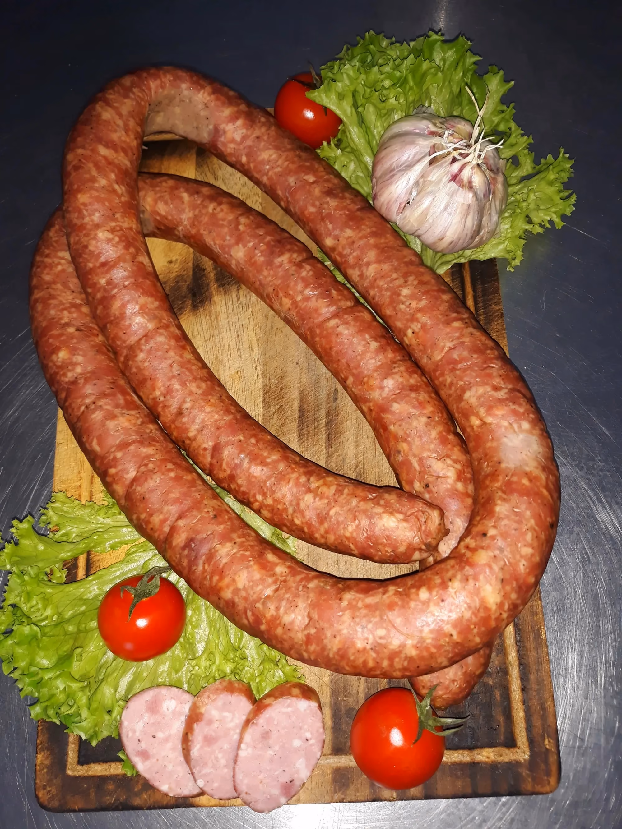 Pętko wiejskiej kiełbasy i kilka plastrów ułożonych na drewnianej desce, udekorowane liśćmi sałaty, pomidorkami koktajlowymi i główką czosnku
