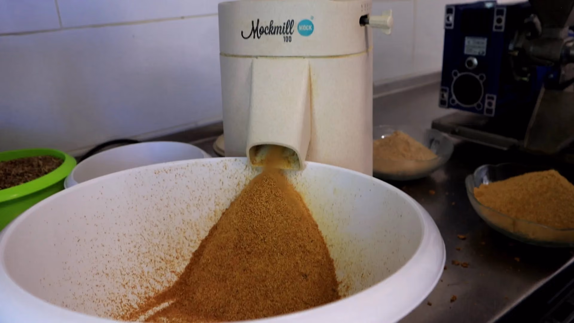 Młynek elektryczny marki Mockmill podczas mielenia zboża do dużej białej miski na tle blatu kuchennego