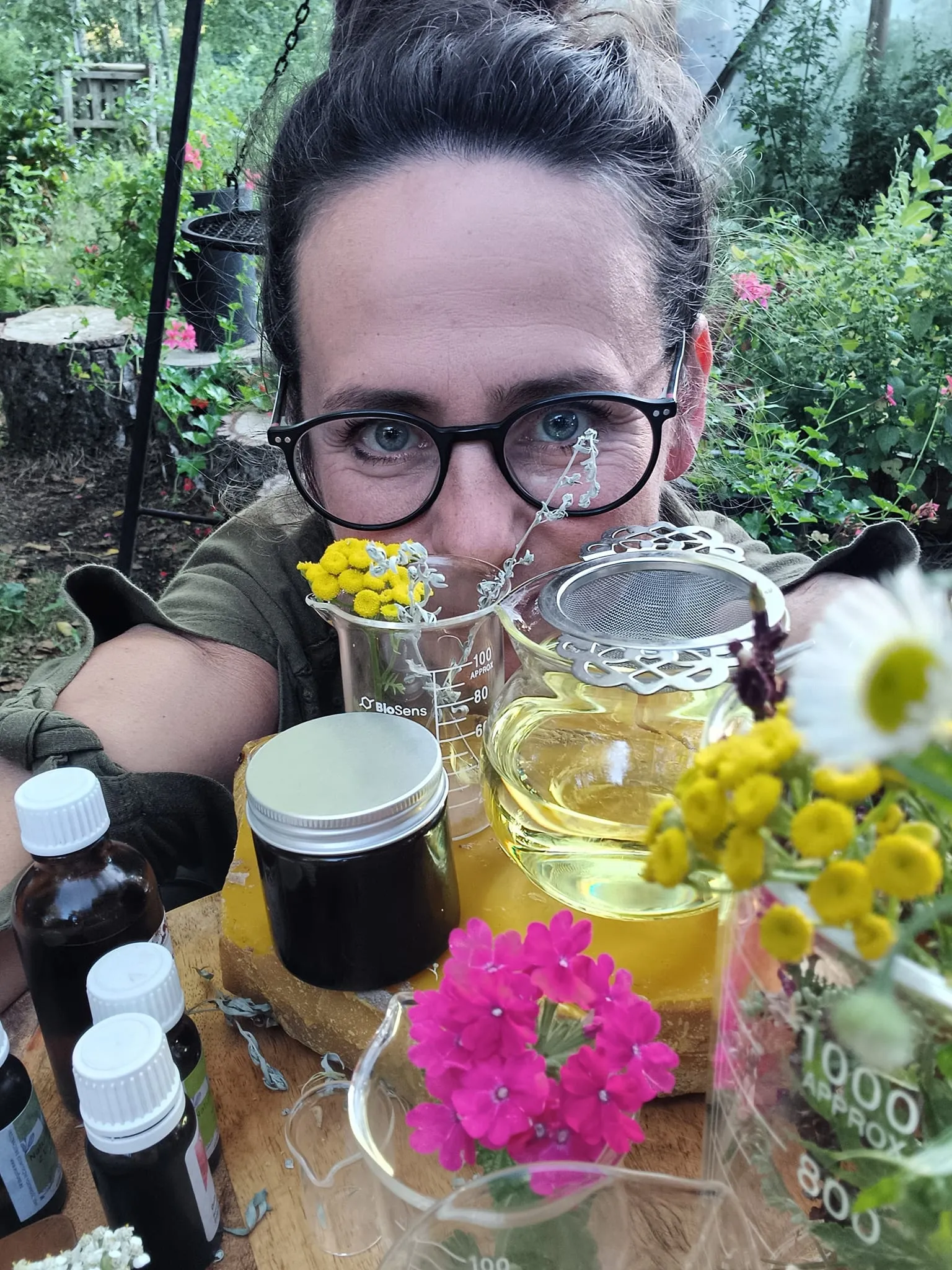 Kobieta w okularach pochyla się nad stołem, na którym znajdują się szklane naczynia laboratoryjne z ziołami, słoiczki z produktami oraz kwitnące kwiaty
