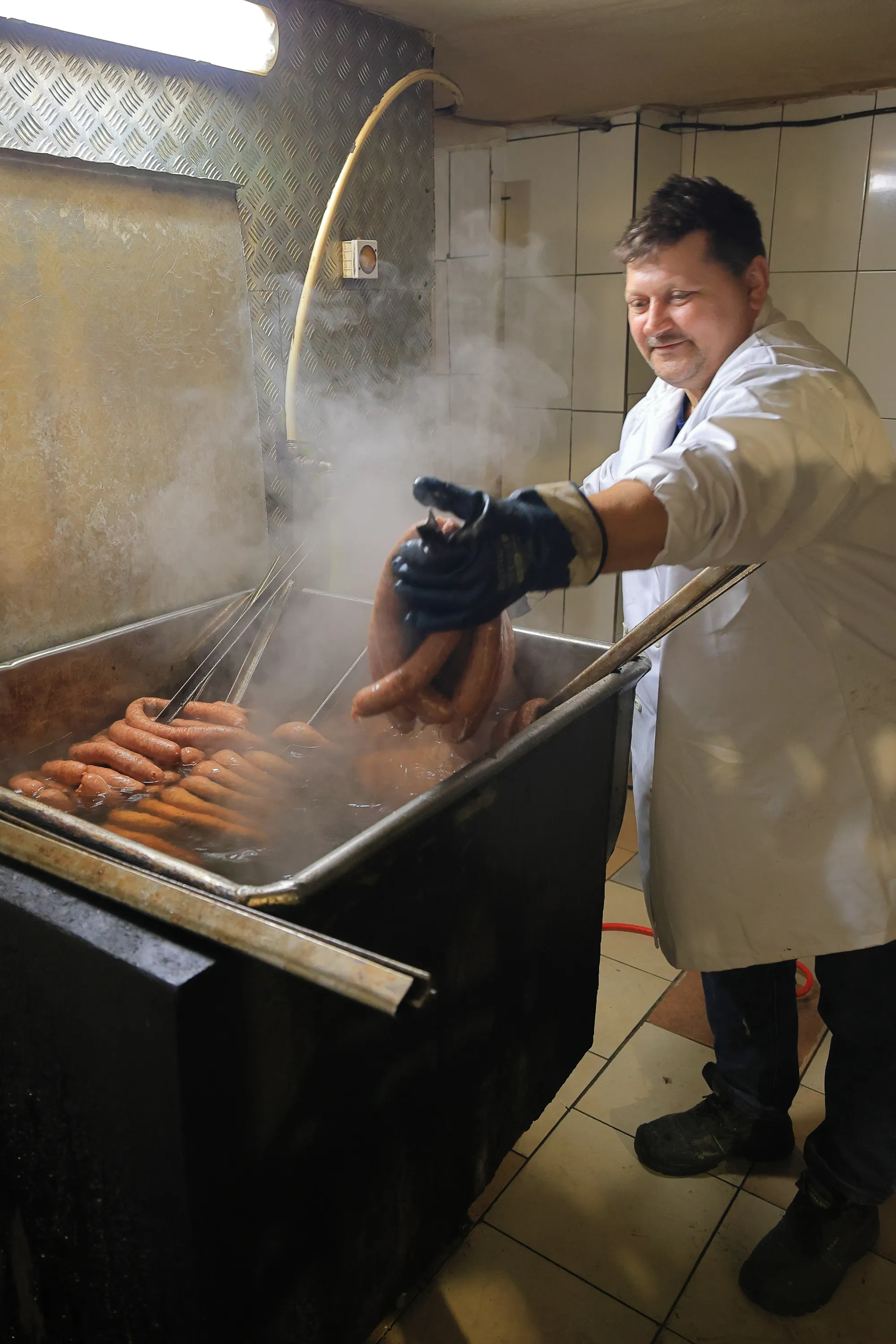 Wędliniarz w białym fartuchu wyjmuje parującą kiełbasę z dużego metalowego zbiornika z gorącą wodą