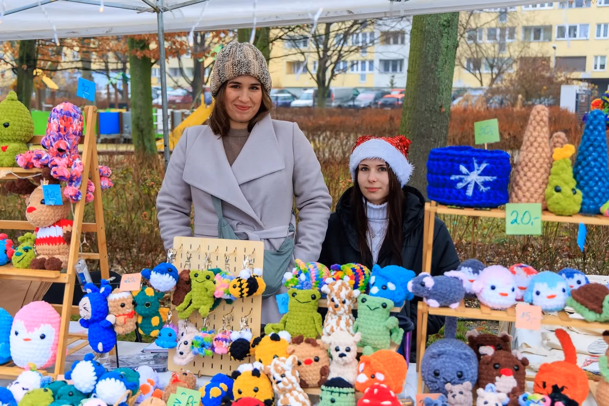 Dwie kobiety stoją za stoiskiem na jarmarku, na którym wyeksponowano bogatą ofertę ręcznie wykonanych, szydełkowanych maskotek w różnych kolorach i kształtach, m.in. zwierząt i breloków.