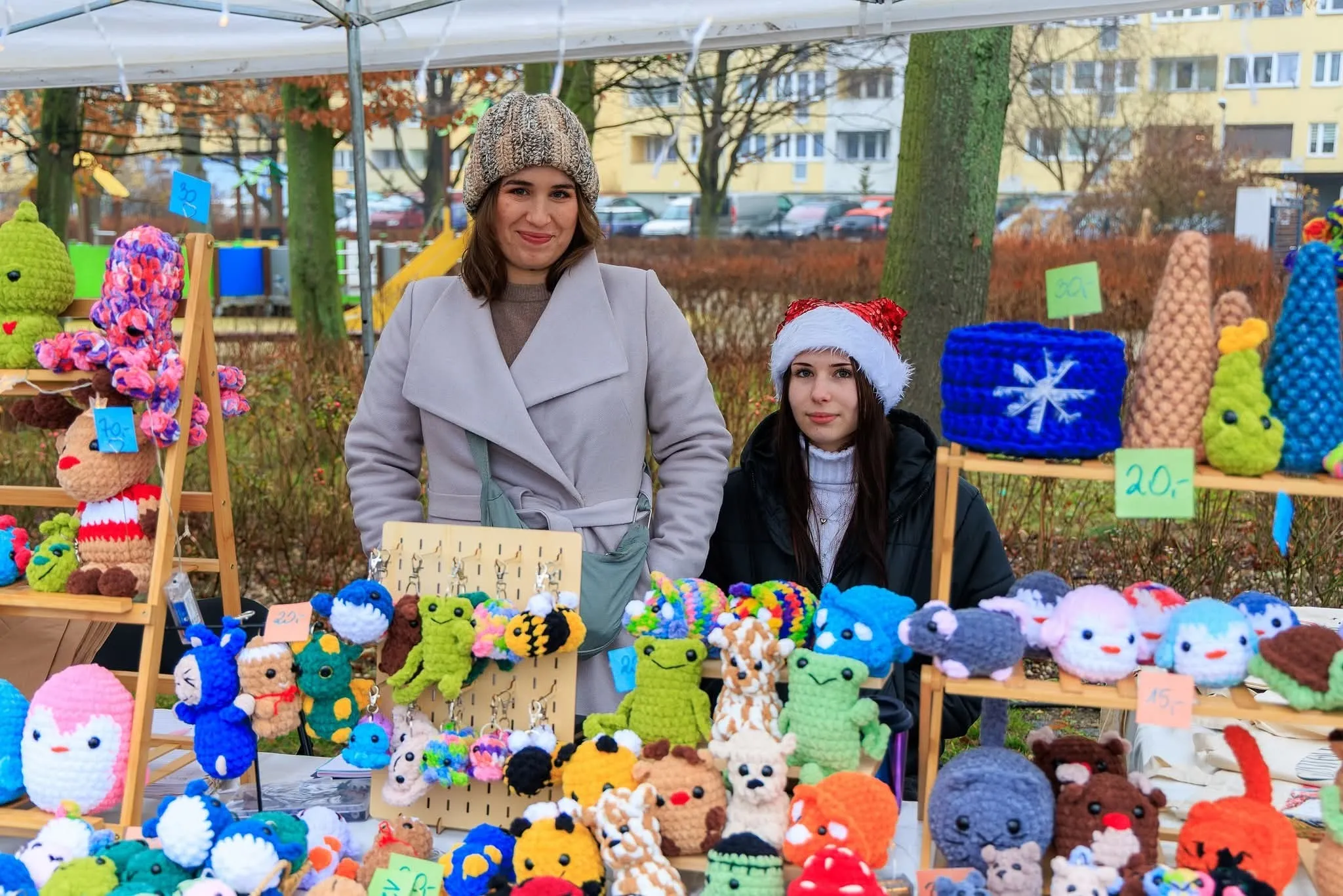 Dwie kobiety stoją za stoiskiem na jarmarku, na którym wyeksponowano bogatą ofertę ręcznie wykonanych, szydełkowanych maskotek w różnych kolorach i kształtach, m.in. zwierząt i breloków.