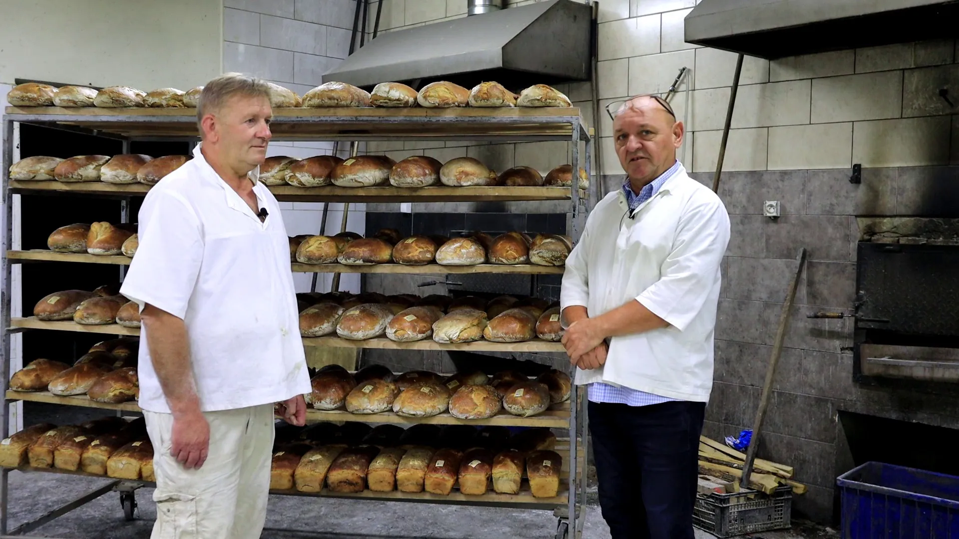 Dwaj mężczyźni w jasnych fartuchach stoją we wnętrzu piekarni obok wysokiego, metalowego regału na kółkach, całkowicie zapełnionego bochenkami chleba. Obok nich widoczny jest piec.