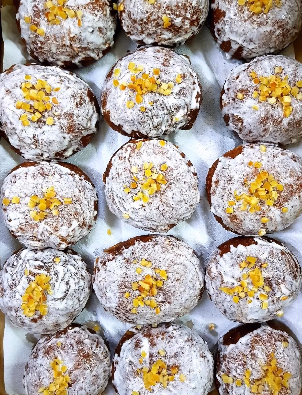 Zbliżenie na pączki z lukrem, ułożone w rzędach na białym papierze. Każdy pączek jest posypany kawałkami kandyzowanej skórki pomarańczowej.