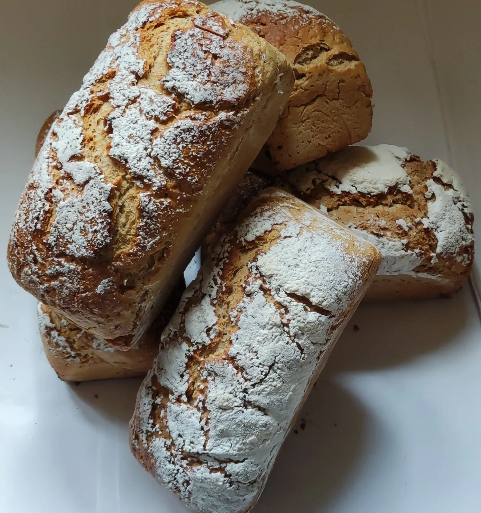 Kilka upieczonych, prostokątnych chlebów foremkowych ułożonych w stos na jasnym tle. Bochenki mają mocno przypieczoną, spękaną skórkę, a ich wierzch jest obficie oprószony mąką.