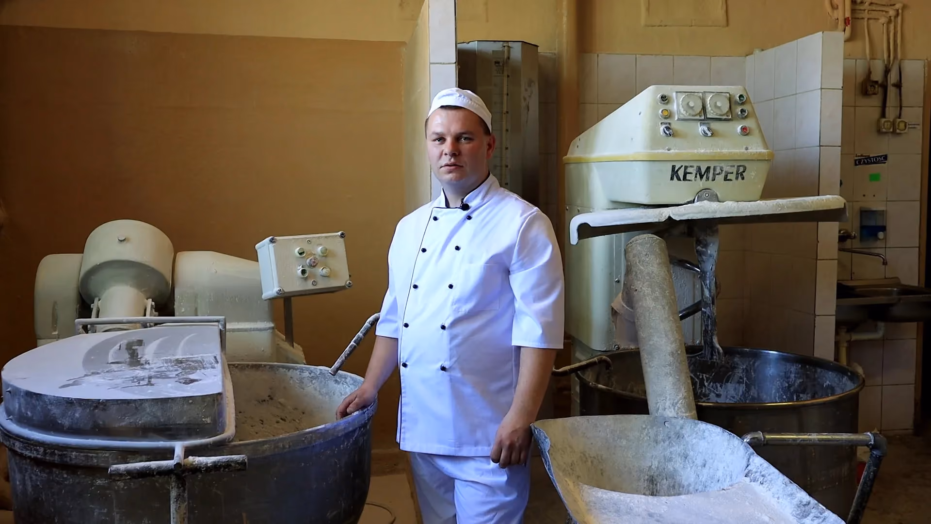 Piekarz w białym kitlu i czapce stoi pośrodku pomieszczenia produkcyjnego, opierając dłoń o krawędź dużej dzieży miksera do ciasta. Po jego prawej stronie stoi druga, wysoka maszyna mieszalna marki Kemper, a całe otoczenie nosi ślady mąki.