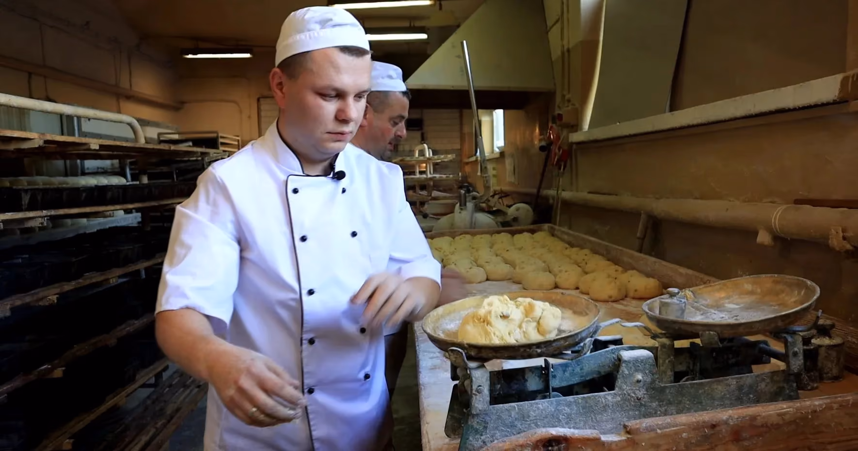 Piekarz w białym kitlu i czapce pracuje przy stole piekarniczym, na którym stoi stara, dwuszalkowa waga mechaniczna z kawałkiem surowego ciasta. W tle, na mąką posypanym blacie, ułożone są uformowane porcje ciasta na chleb, a obok pracuje drugi piekarz.