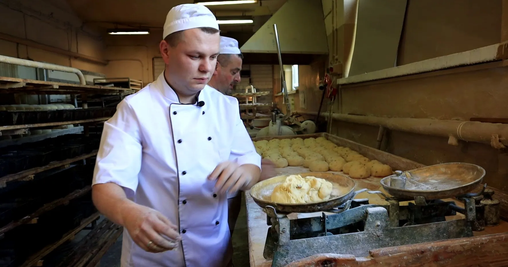 Piekarz w białym kitlu i czapce pracuje przy stole piekarniczym, na którym stoi stara, dwuszalkowa waga mechaniczna z kawałkiem surowego ciasta. W tle, na mąką posypanym blacie, ułożone są uformowane porcje ciasta na chleb, a obok pracuje drugi piekarz.