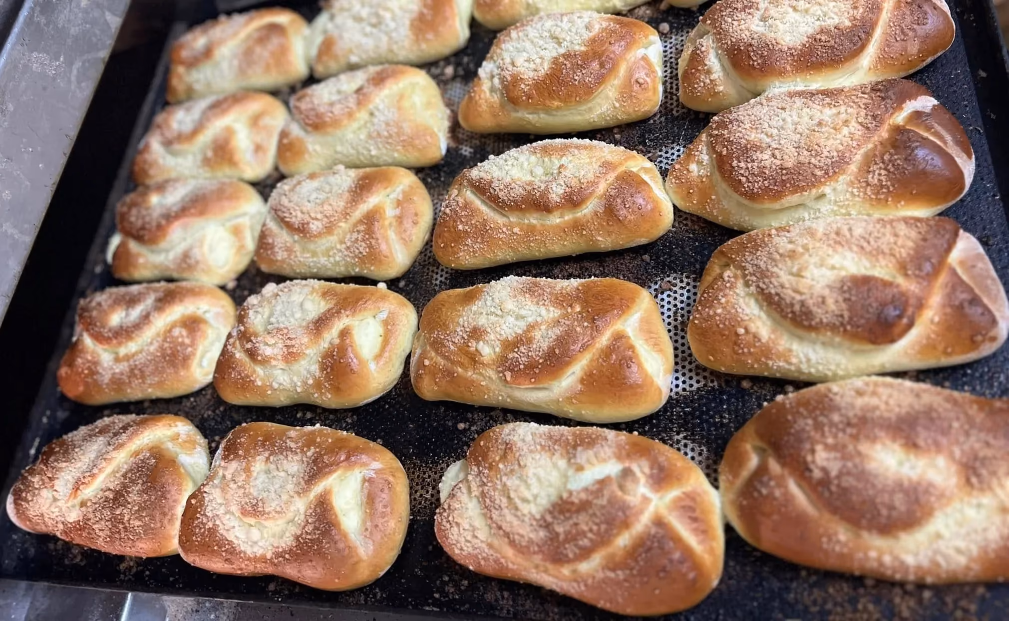 Upieczone, podłużne drożdżówki o błyszczącej, rumianej skórce, ułożone w rzędach na dużej, czarnej blasze piekarniczej. Część z nich jest posypana jasną kruszonką.