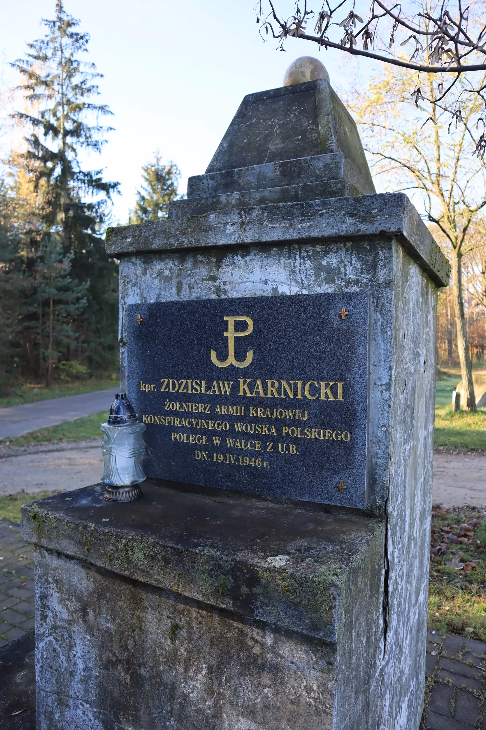 Widok całego pomnika z betonowym cokołem zwieńczonym kulą. Na froncie znajduje się tablica poświęcona kapralowi Januszowi „Newadzie”, żołnierzowi Armii Krajowej poległemu w 1945 roku. U podstawy ustawiono znicze. W tle widoczne są pola i domy