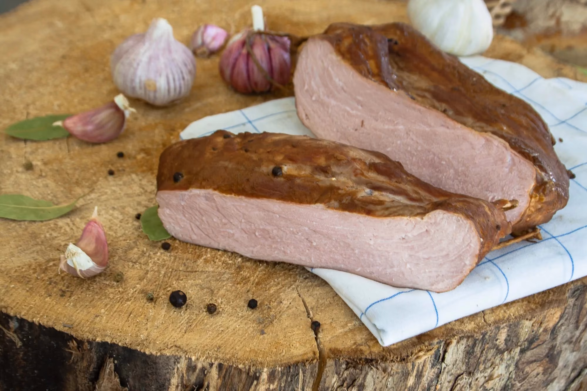 Dwa duże, przekrojone wzdłuż kawałki wędzonej szynki z widocznym ziarnistym pieprzem na ciemnej skórce, ułożone na białej ściereczce w niebieską kratę. Całość leży na drewnianym pniu w otoczeniu główek i ząbków czosnku oraz liści laurowych.