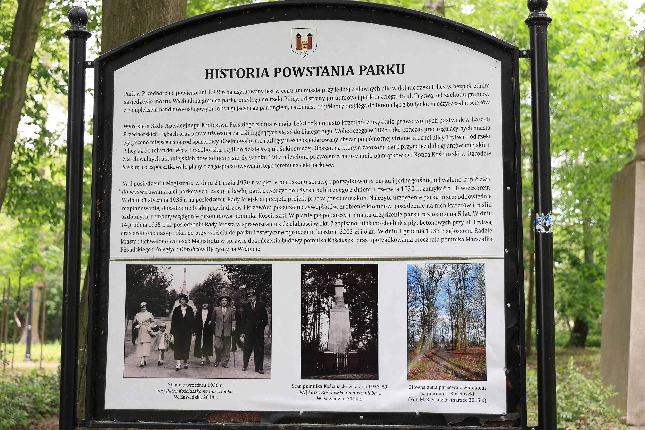 Na zdjęciu znajduje się duża tablica edukacyjna ustawiona na terenie parku. Tablica zatytułowana „Historia powstania parku” zawiera tekst opisujący dzieje założenia i rozwoju parku miejskiego oraz kilka fotografii archiwalnych i współczesnych parku
