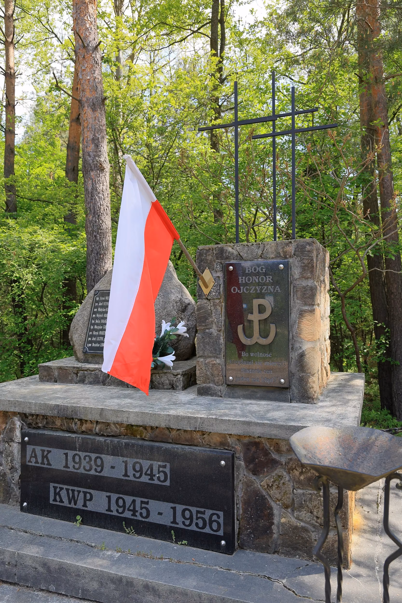 Na zdjęciu przedstawiono boczny widok pomnika poświęconego żołnierzom Armii Krajowej i Konspiracyjnego Wojska Polskiego. Widoczna jest biało-czerwona flaga, tablica z symbolem Polski Walczącej oraz napisem „Bóg, Honor, Ojczyzna”