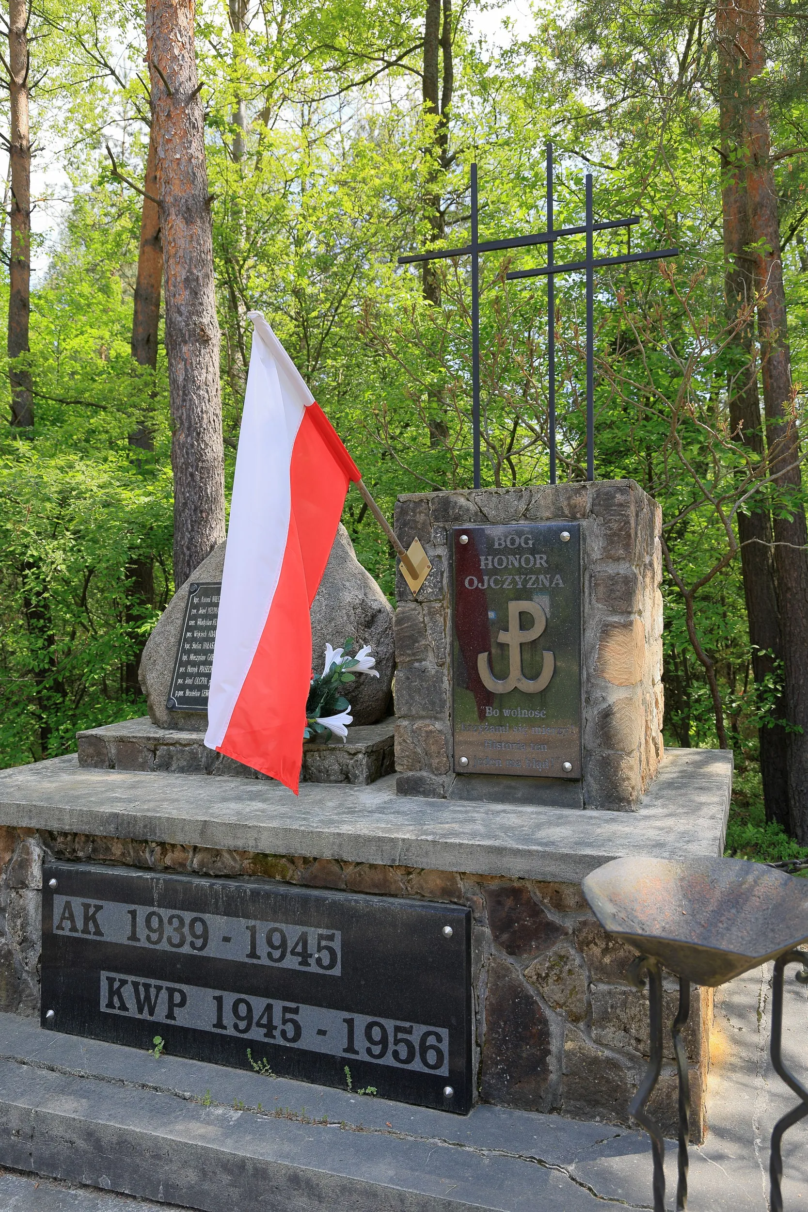 Na zdjęciu przedstawiono boczny widok pomnika poświęconego żołnierzom Armii Krajowej i Konspiracyjnego Wojska Polskiego. Widoczna jest biało-czerwona flaga, tablica z symbolem Polski Walczącej oraz napisem „Bóg, Honor, Ojczyzna”