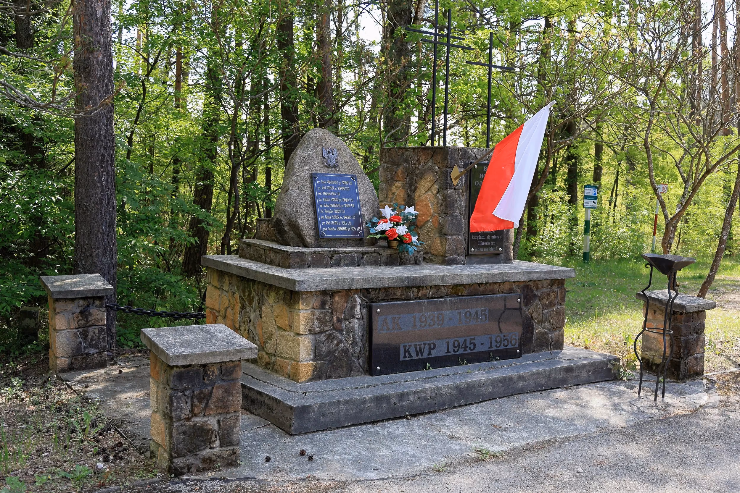 Na zdjęciu widoczny jest kamienny pomnik pamięci usytuowany wśród drzew i zieleni. Monument składa się z kamiennej podstawy, głazu z tablicą pamiątkową oraz pionowego elementu z symbolem Polski Walczącej
