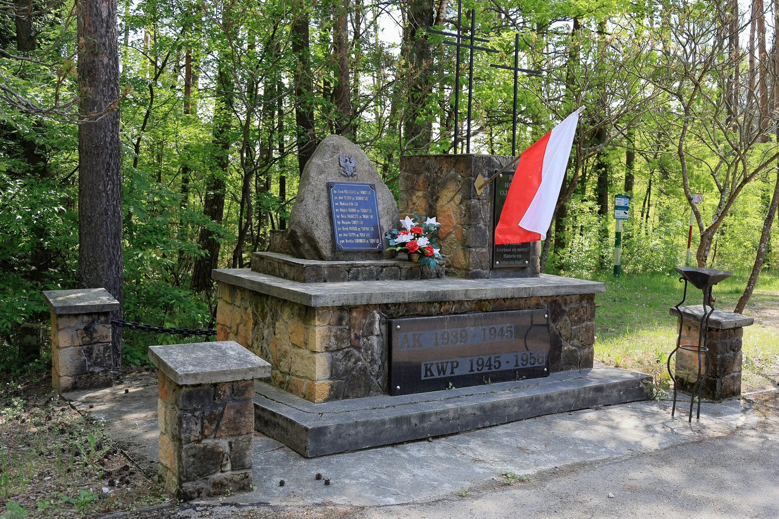 Na zdjęciu widoczny jest kamienny pomnik pamięci usytuowany wśród drzew i zieleni. Monument składa się z kamiennej podstawy, głazu z tablicą pamiątkową oraz pionowego elementu z symbolem Polski Walczącej