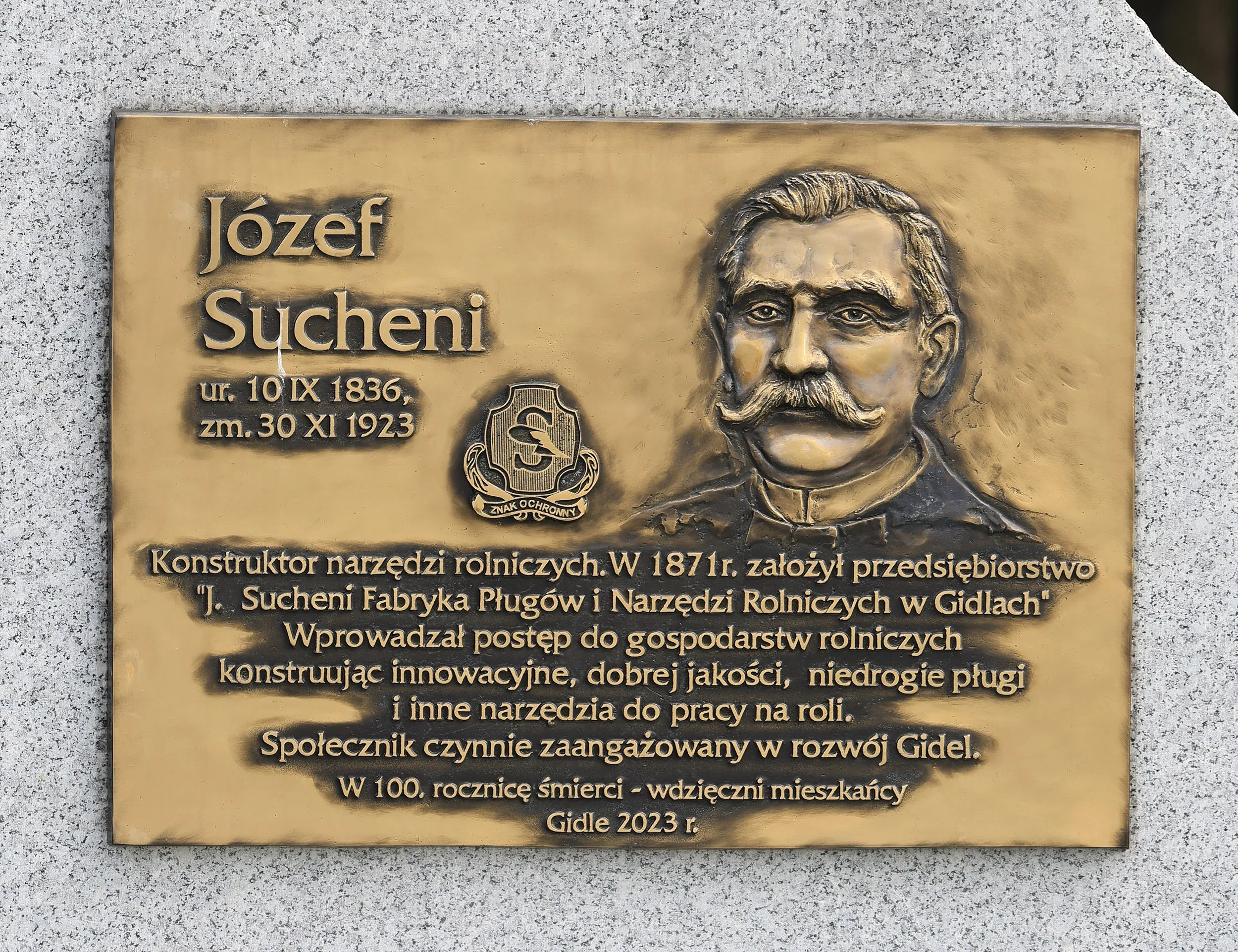 Tablica pamiątkowa poświęcona Józefowi Sucheniowi, konstruktorowi narzędzi rolniczych i założycielowi fabryki pługów w Gidlach, z płaskorzeźbą jego portretu.