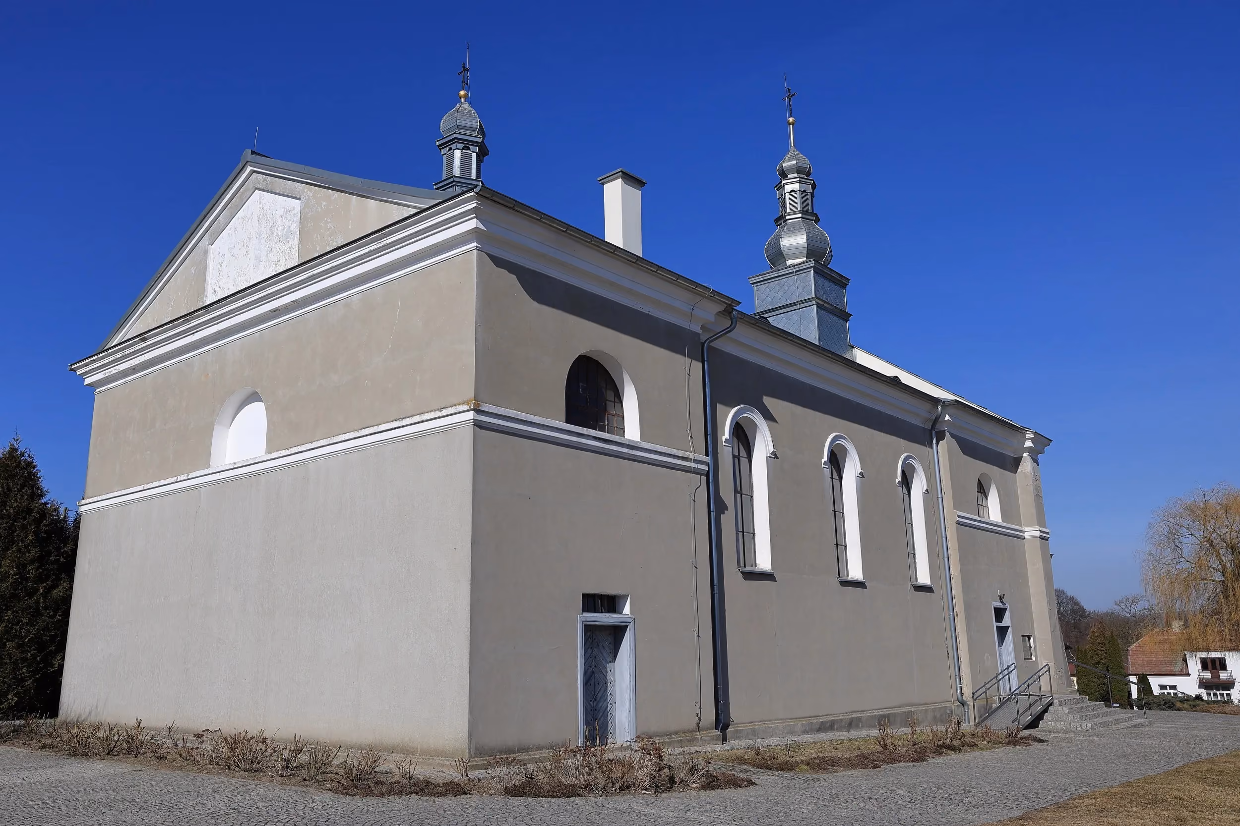 Widok bocznej elewacji kościoła z wysokimi oknami i dwiema wieżyczkami. Jasny budynek stoi przy brukowanym placu w słoneczny dzień.