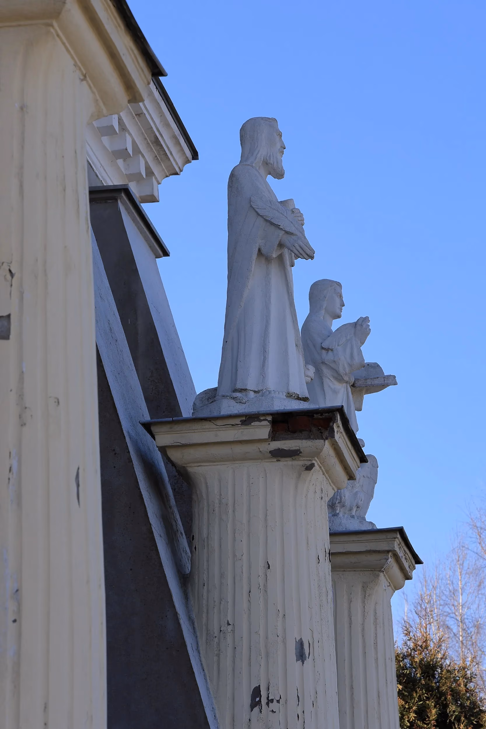 Detal elewacji kościoła z białymi figurami świętych ustawionymi na kolumnach przy fasadzie. Rzeźby widoczne na tle bezchmurnego, błękitnego nieba.