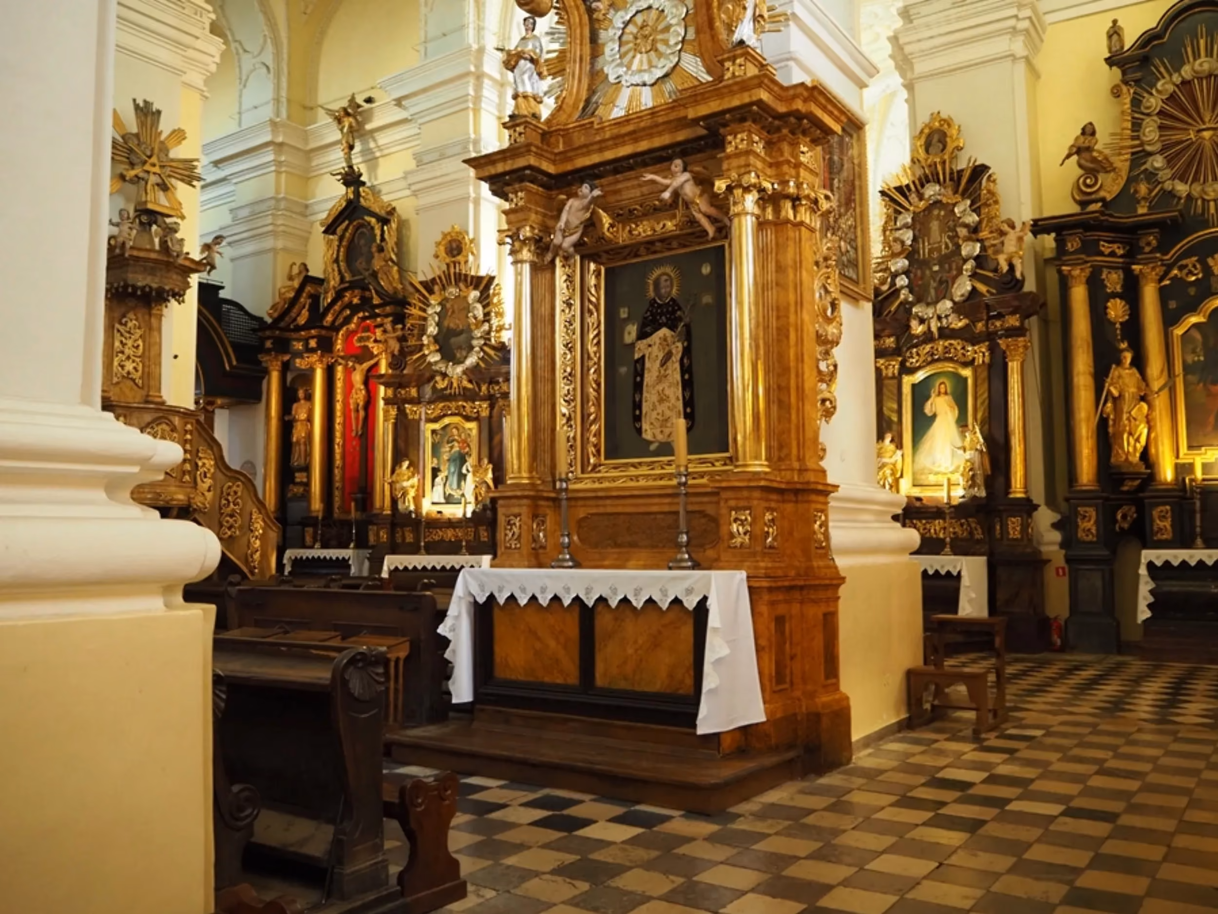 Wnętrze bazyliki z bocznym ołtarzem i bogato zdobionymi, barokowymi nastawami. Widoczne ławki, biało-czarna posadzka oraz obrazy religijne w złoconych ramach.